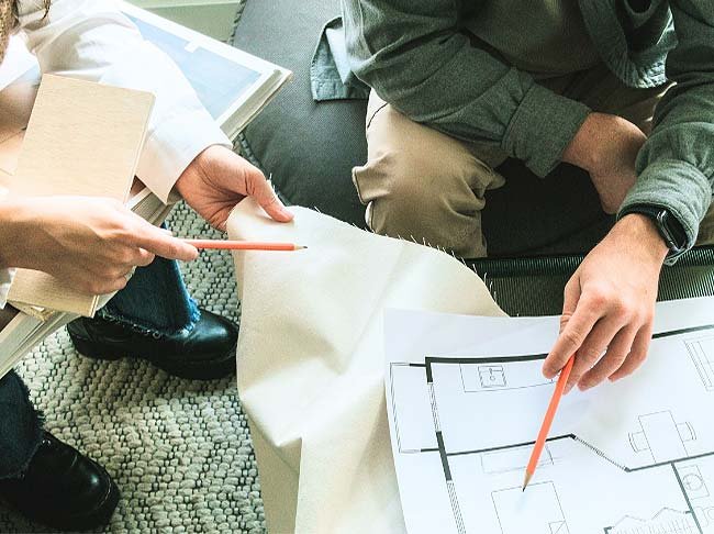 Laps of two people sitting at a table, one holding fabric the other pointing at design plans with a pencil. 