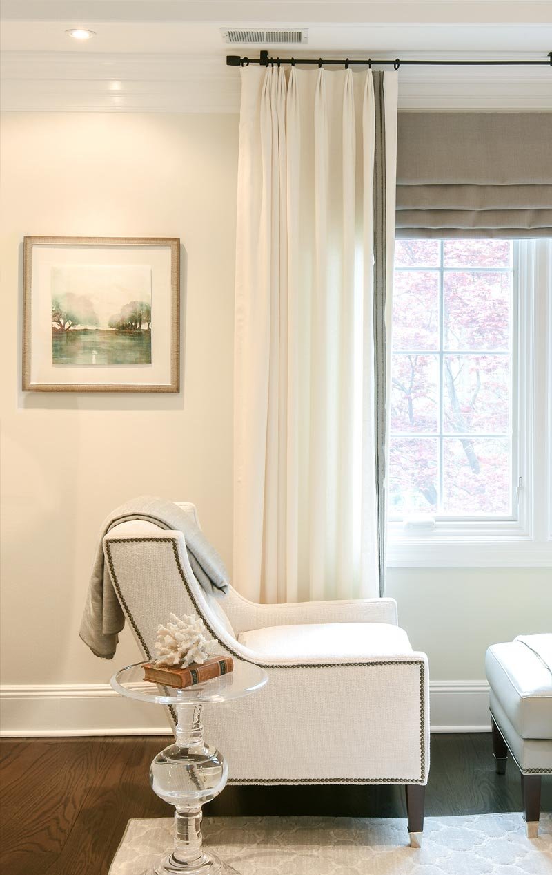 Side view of an elegant white chair with dark brown trim. Glass end table next to the chair. Behind is a window looking out to a blossoming tree.